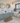 A bright, modern bathroom featuring a light blue double-sink vanity with white ceramic vessel sinks, a white freestanding bathtub with a draped towel, patterned gray and white floor tiles, and a brown coir bath mat.