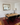 A rustic wooden dining table with a black metal frame and six black chairs, set on a red patterned area rug in a dining room with white wainscoting and a large gold-framed mirror.